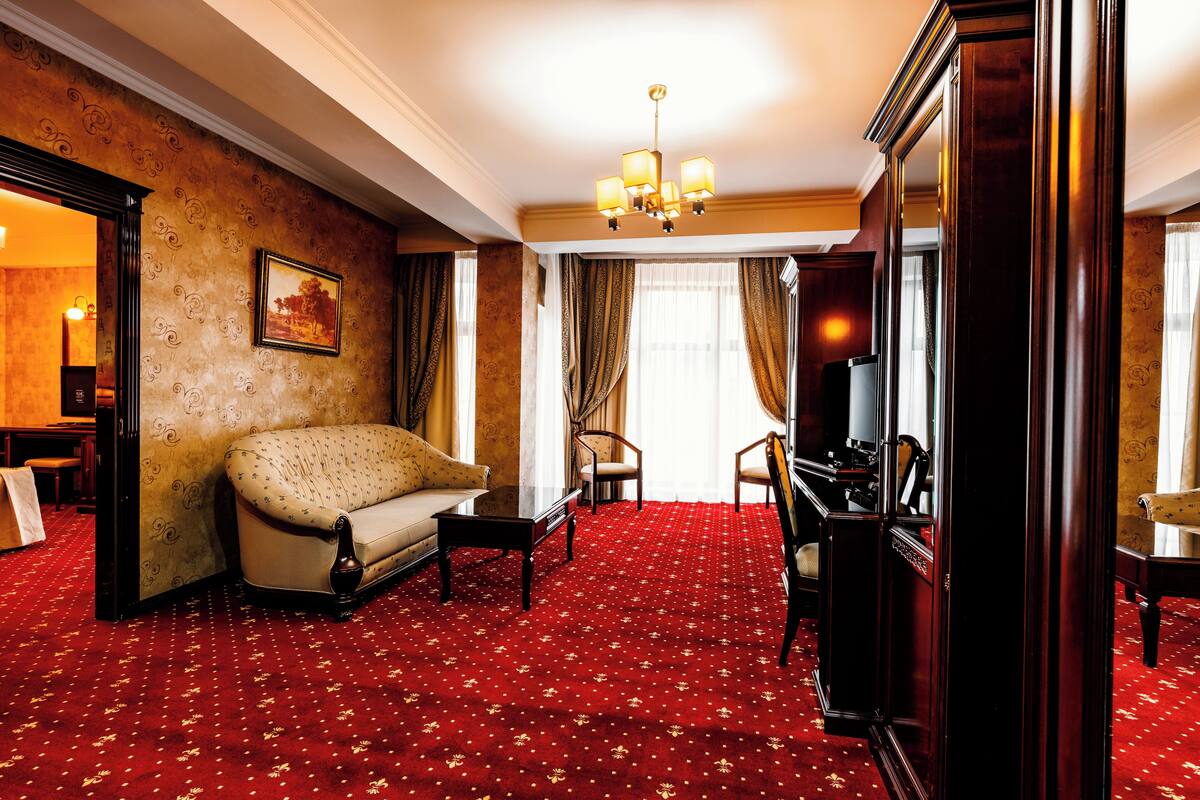 A hotel room with a red carpet, a sofa, a chair, a television, and a painting on the wall.
