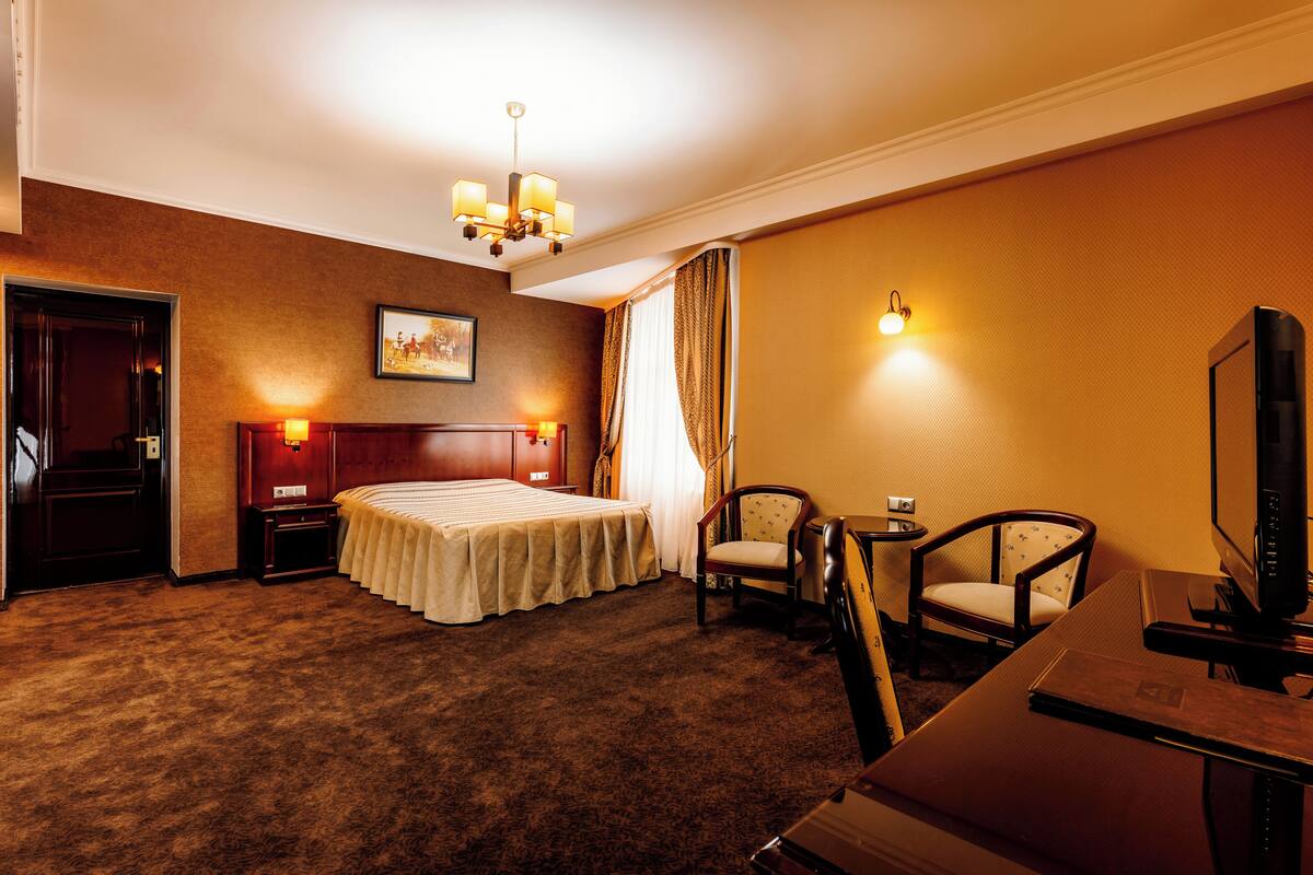 A hotel room with a bed, two chairs, a desk, a television, and a chandelier.