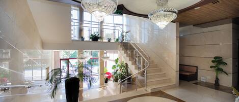 A spacious lobby with a grand staircase, large chandeliers, and potted plants.