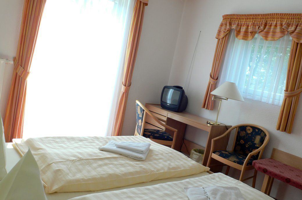 Ein Hotelzimmer mit einem Bett, einem Schreibtisch mit Fernseher, einem Sessel und einem Fenster mit Vorhängen.
