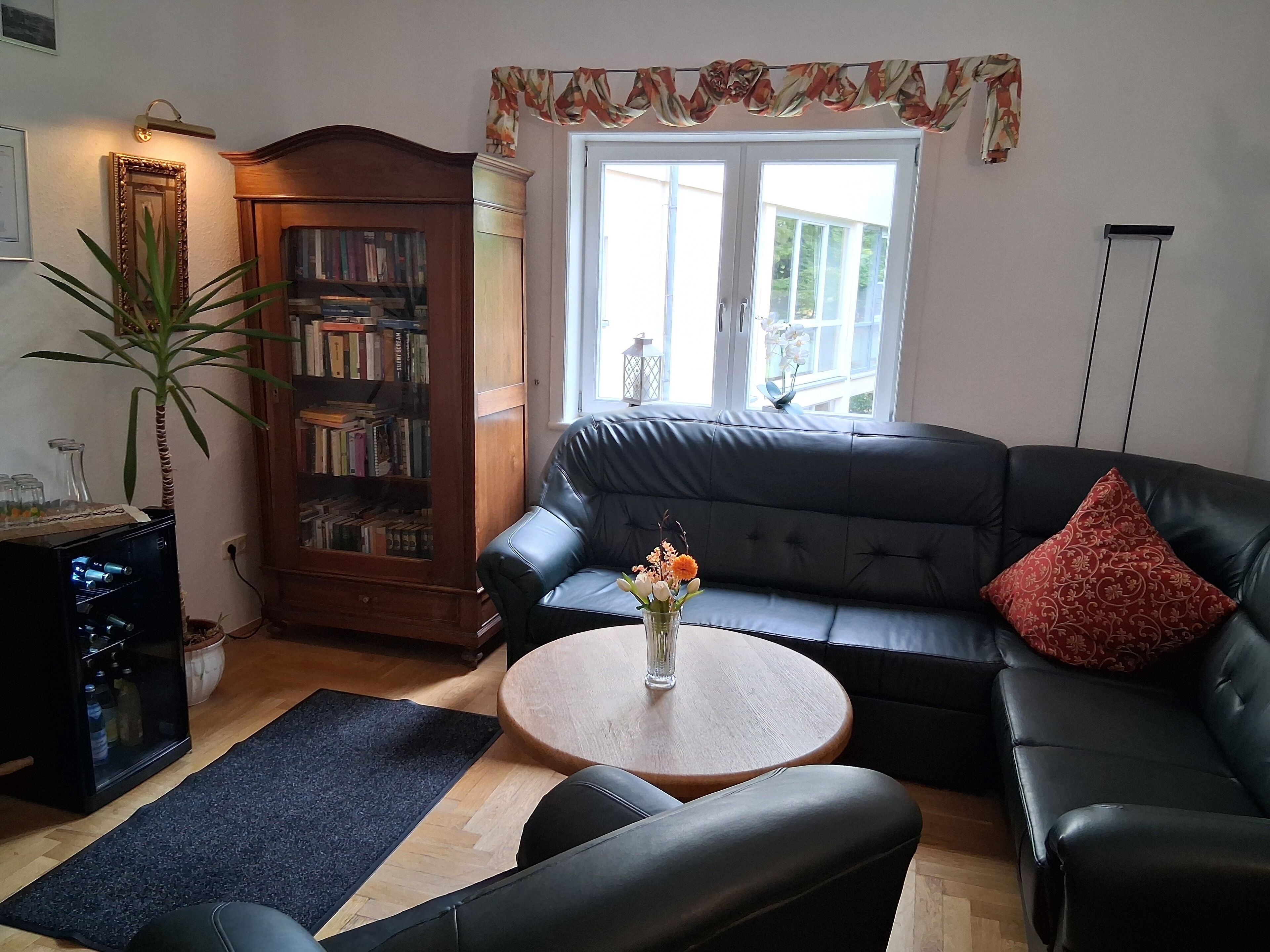 A living room with a leather sofa, a small round table with a vase of flowers, a bookshelf, and a large window.