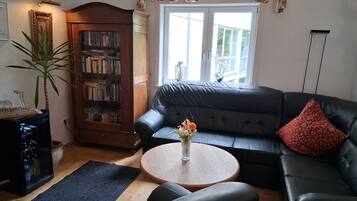 A living room with a leather sofa, a small round table with a vase of flowers, a bookshelf, and a large window.