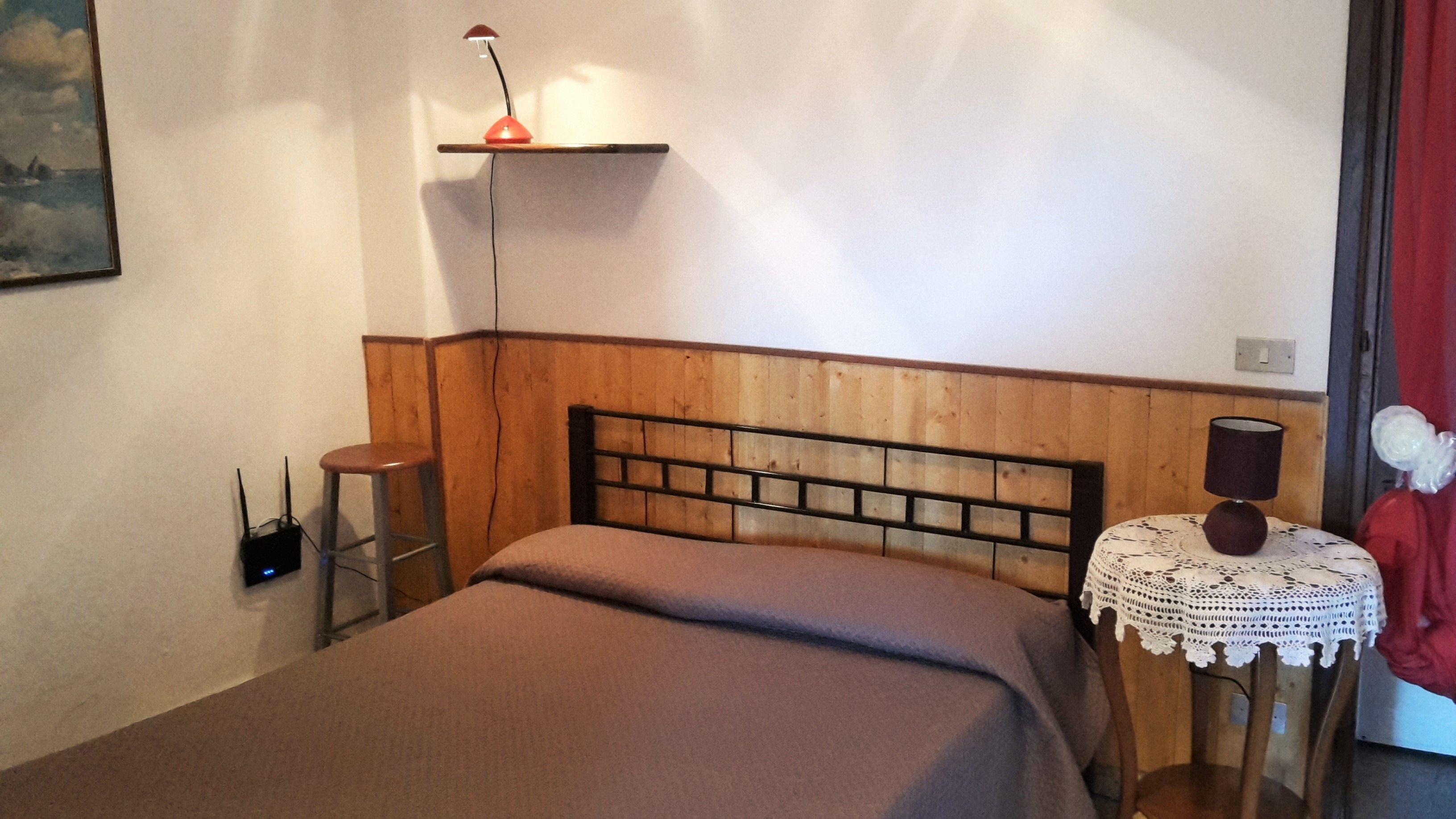 Une chambre à coucher avec une tête de lit en bois, une table de chevet avec une lampe, une chaise rouge et une petite table recouverte d’une nappe en dentelle.