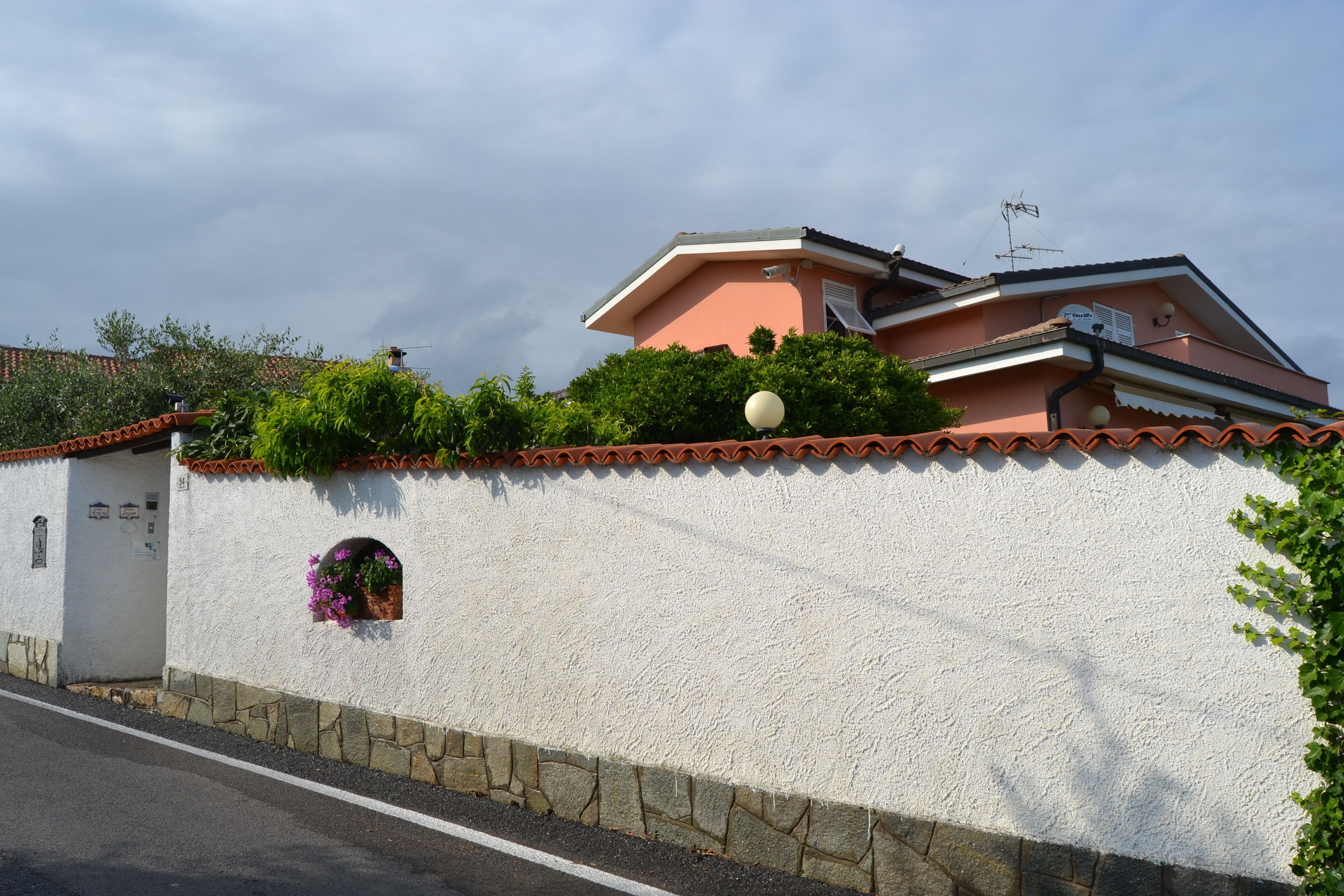 A white stucco wall with a stone base, a small window with a flower box, and a street in front.