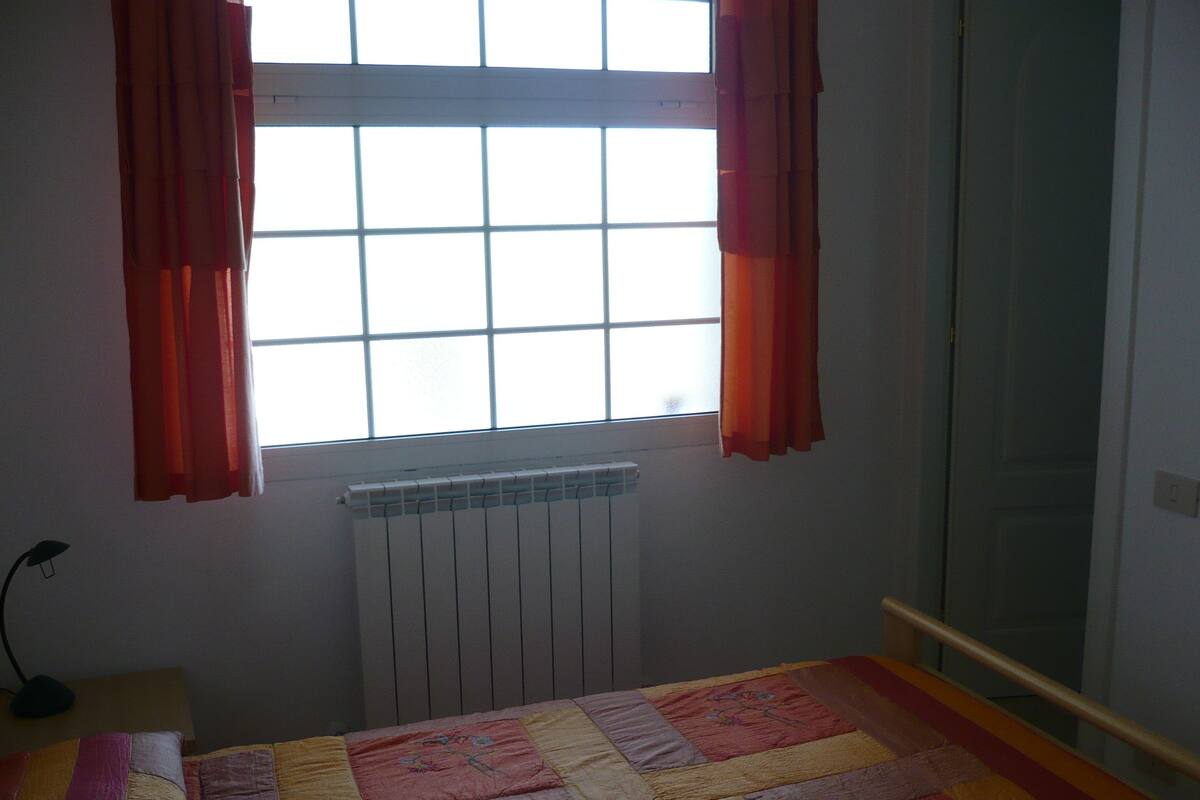 A bedroom with a bed, a window with curtains, a radiator, and a door.