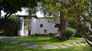 A garden with a white building, stone pathway, and a tree with hanging planters. - Azienda Agrituristica Villa Arianna (Imperia)