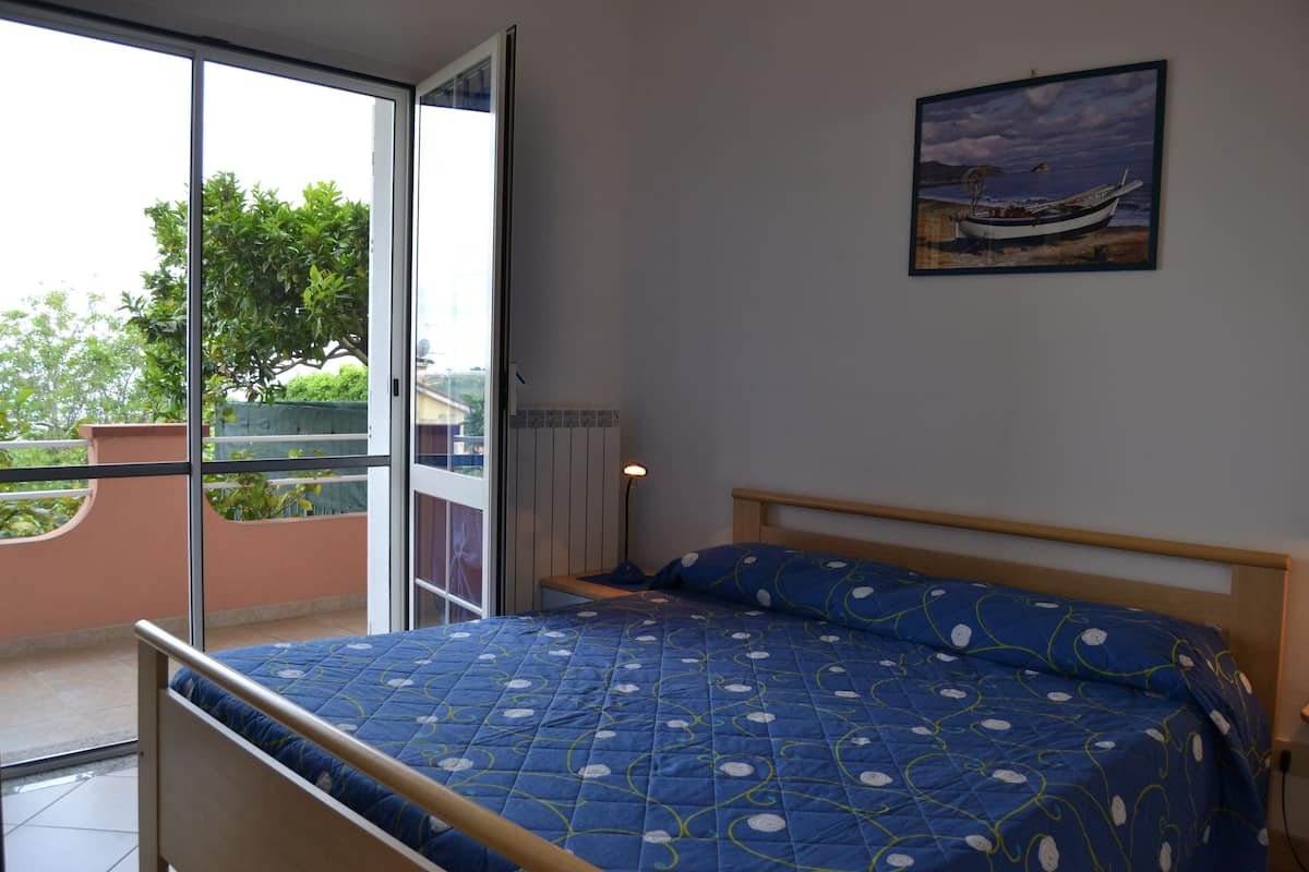 A bedroom with a bed, a painting of a boat on the wall, and a balcony with a view of greenery and buildings.