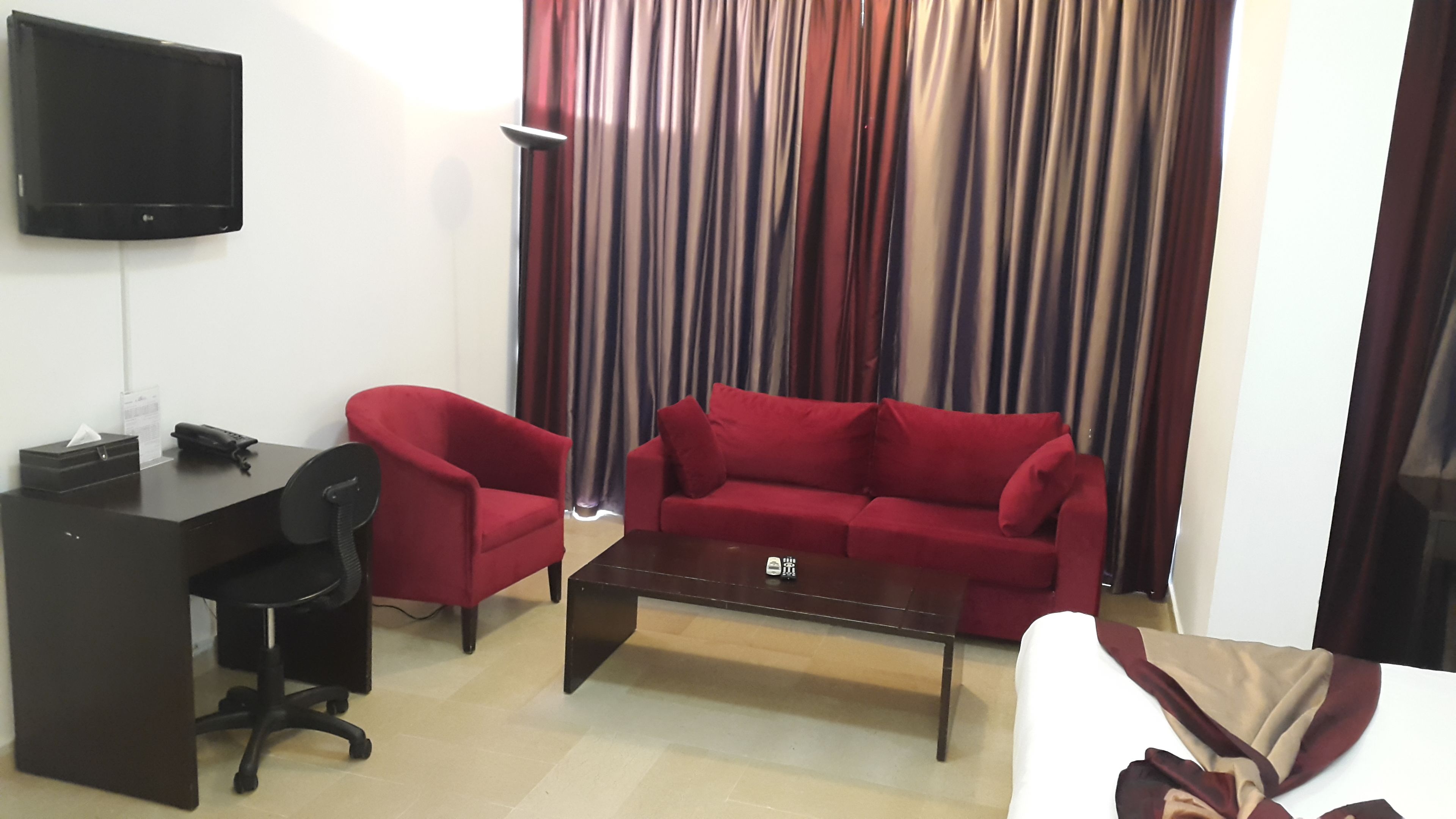 A living room with a red sofa, a black office chair, a coffee table, a TV mounted on the wall, and a curtain with a striped pattern.