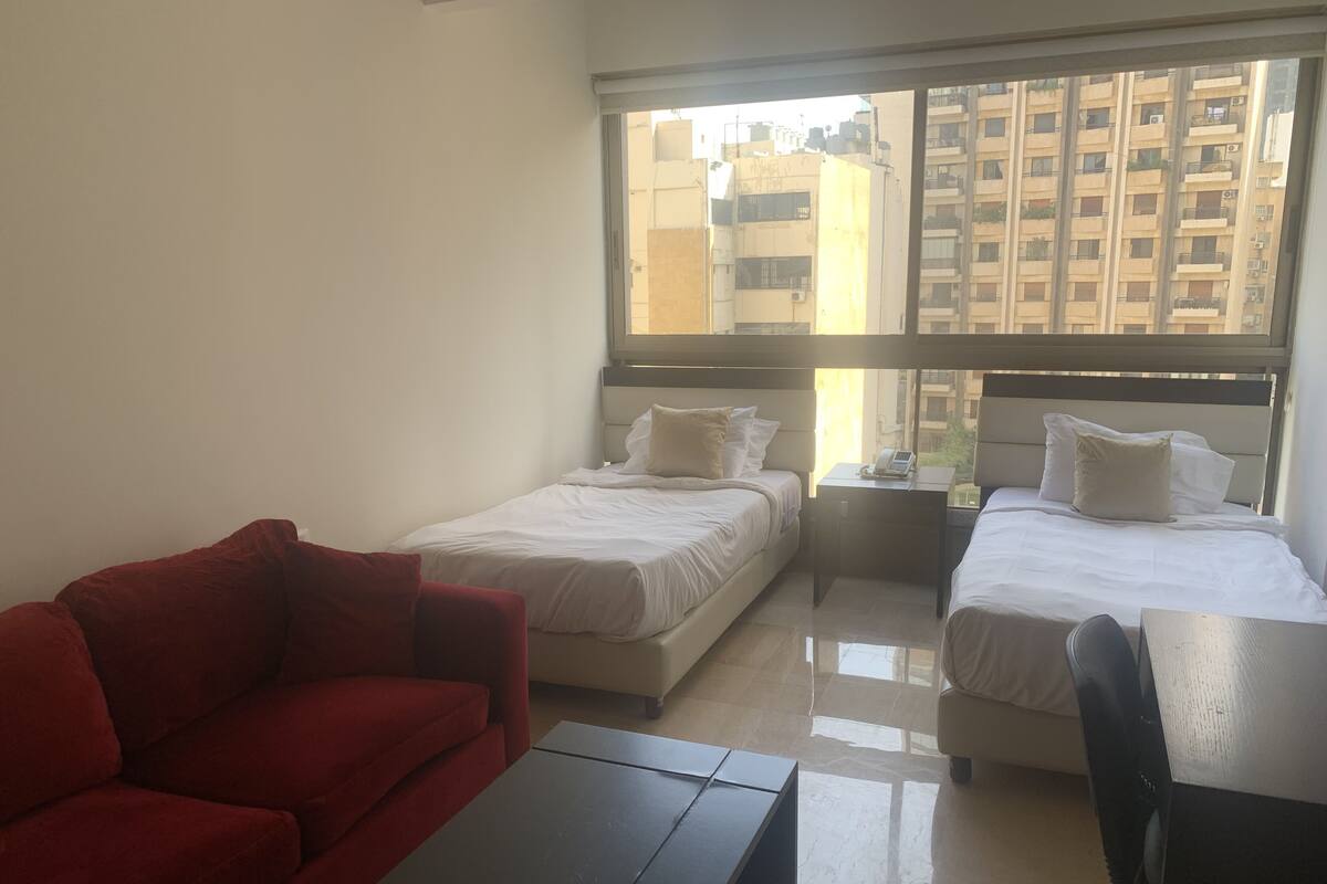 A hotel room with two beds, a red sofa, a small table, and a window with a city view.