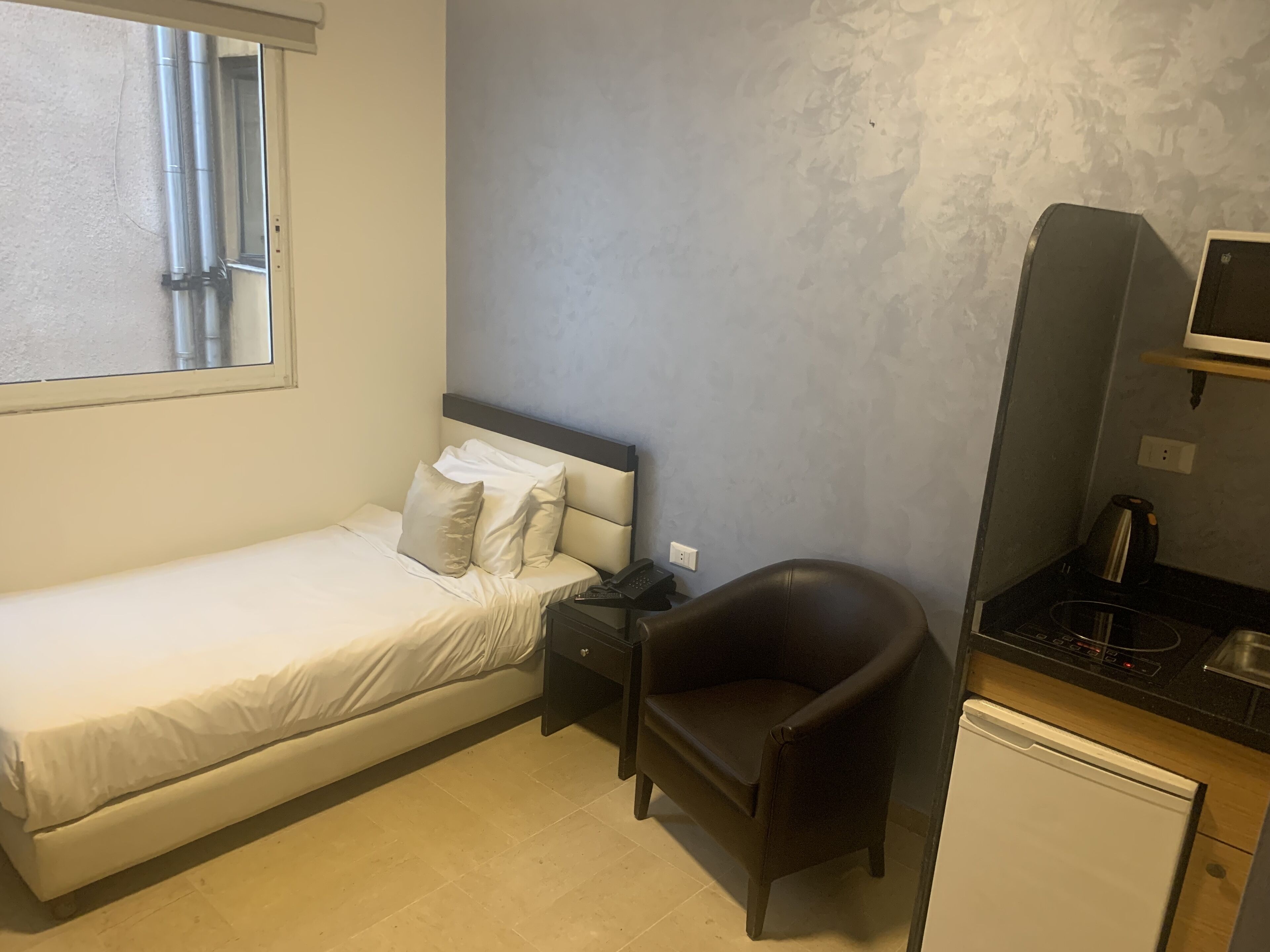 A small, neatly arranged bedroom with a single bed, a chair, a small desk, and a compact kitchenette.