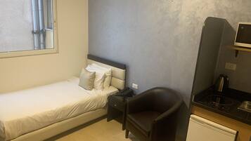 A small, neatly arranged bedroom with a single bed, a chair, a small desk, and a compact kitchenette.