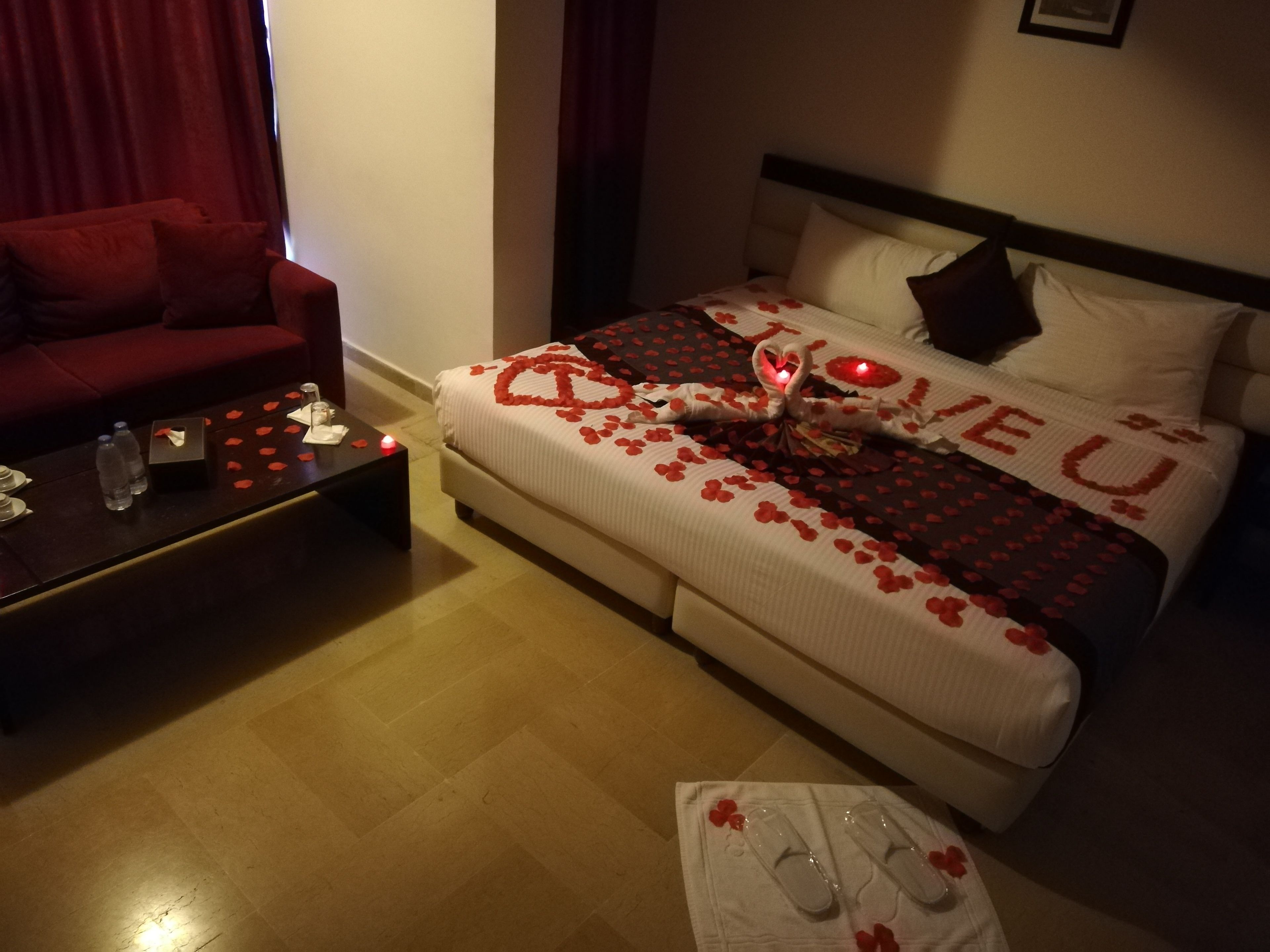 Ein Hotelzimmer mit einem Bett, das mit Rosenblüten und einem herzförmigen Schild mit der Aufschrift 'Ich liebe dich' dekoriert ist, einem roten Sofa, einem Couchtisch mit einer Flasche und Gläsern sowie einem kleinen Tisch mit Hausschuhen.