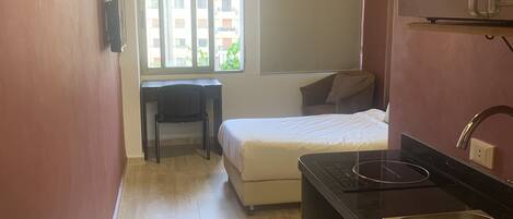 A compact room with a kitchenette, a bed, and a window with a view of buildings.
