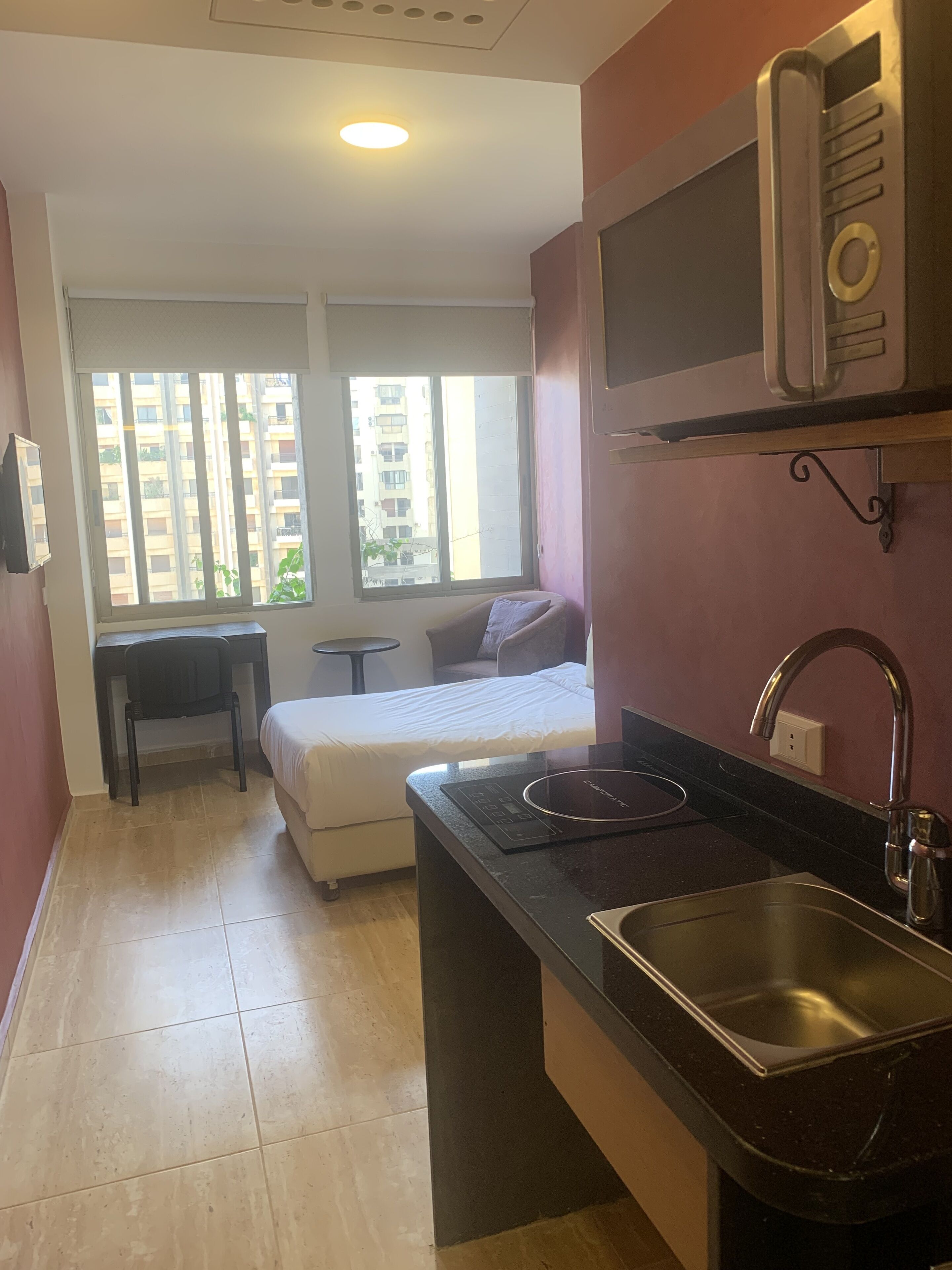 A compact living space with a kitchenette, a bed, a desk, and a window with a view of a building.