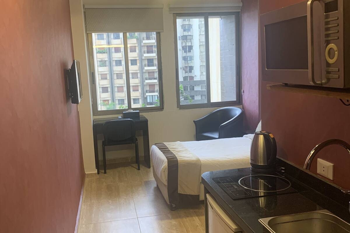A compact hotel room with a kitchenette, a bed, a desk, and large windows offering views of a residential building.