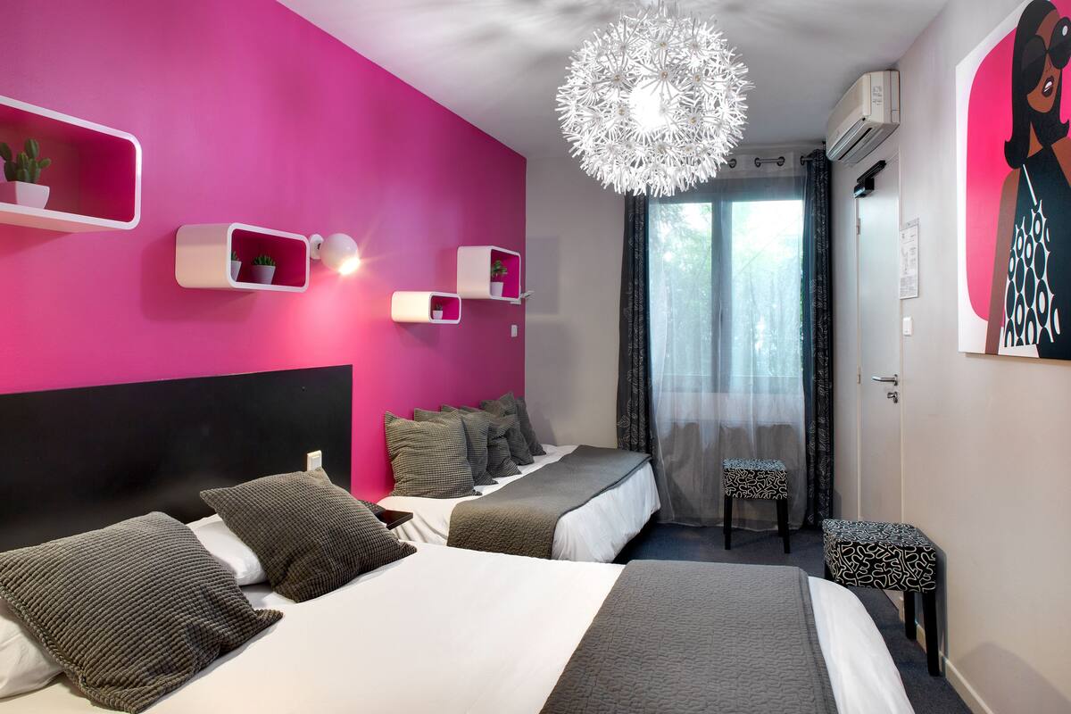 A modern hotel room with a pink accent wall, a large bed with grey bedding, a ceiling light fixture, and a window with sheer curtains.