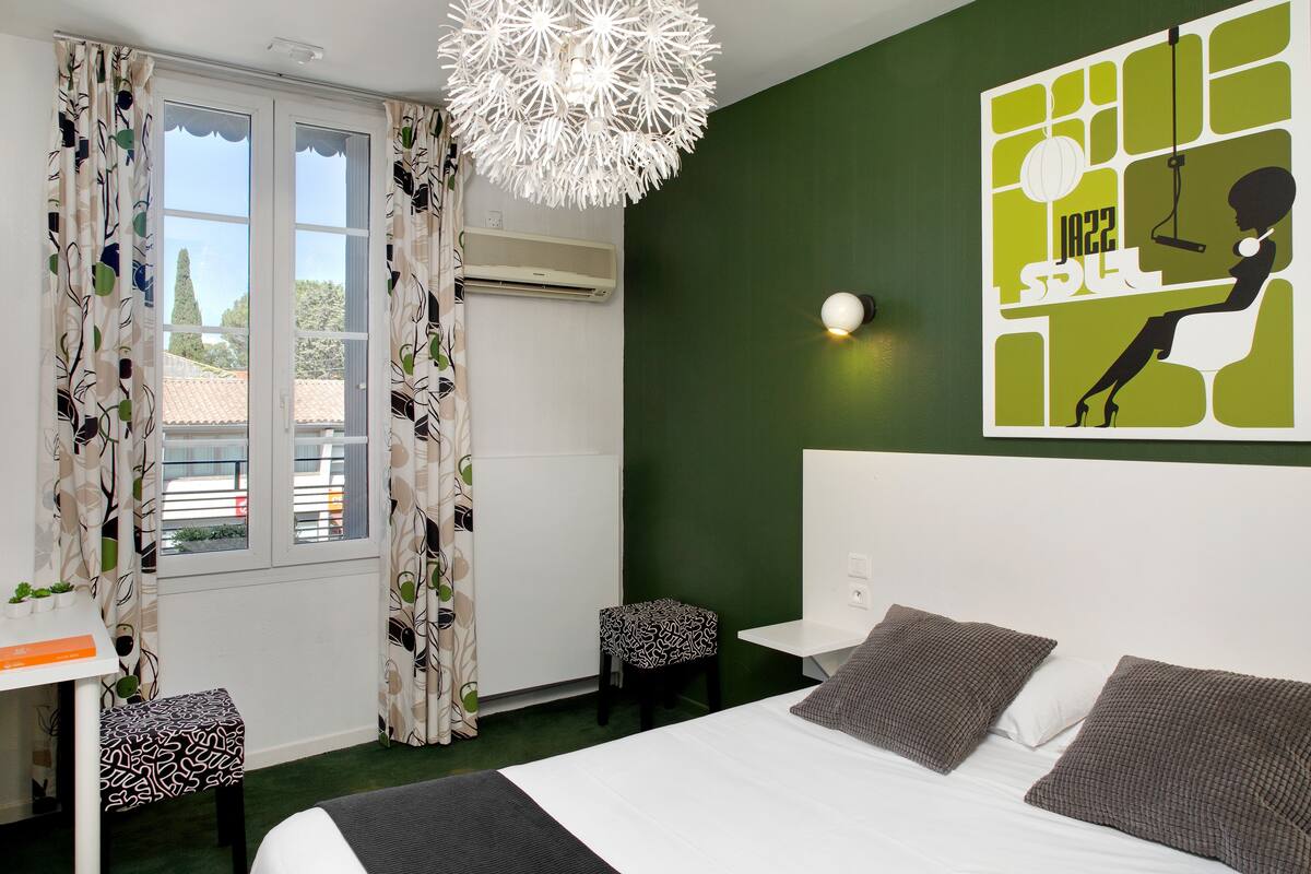 A modern hotel room with a large bed, a desk, and a chair. There is a window with curtains, a wall-mounted air conditioner, and a decorative light fixture.