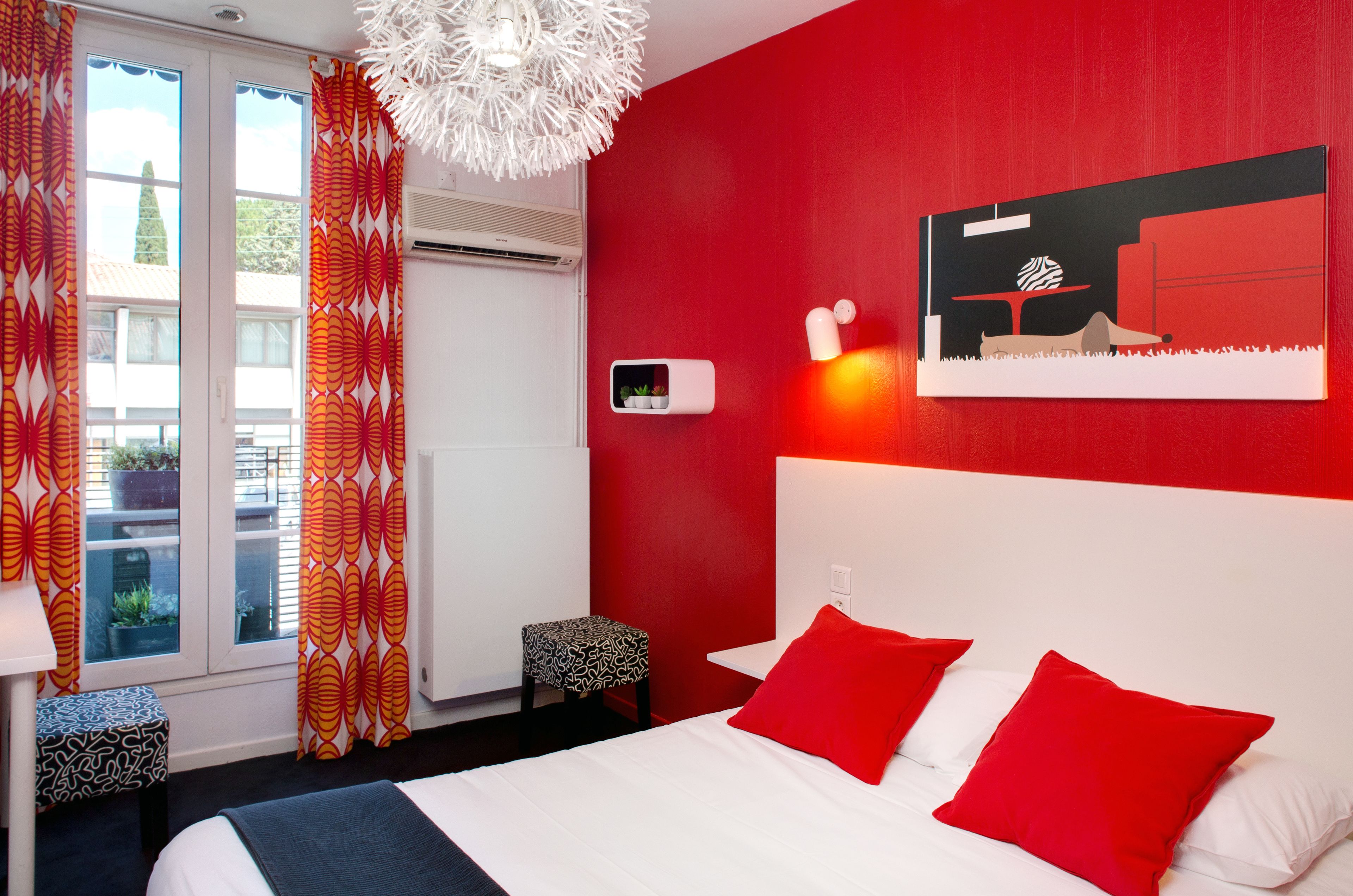 A hotel room with a bed, red pillows, a white headboard, a window with curtains, a wall-mounted air conditioner, and a small bedside table with a lamp.