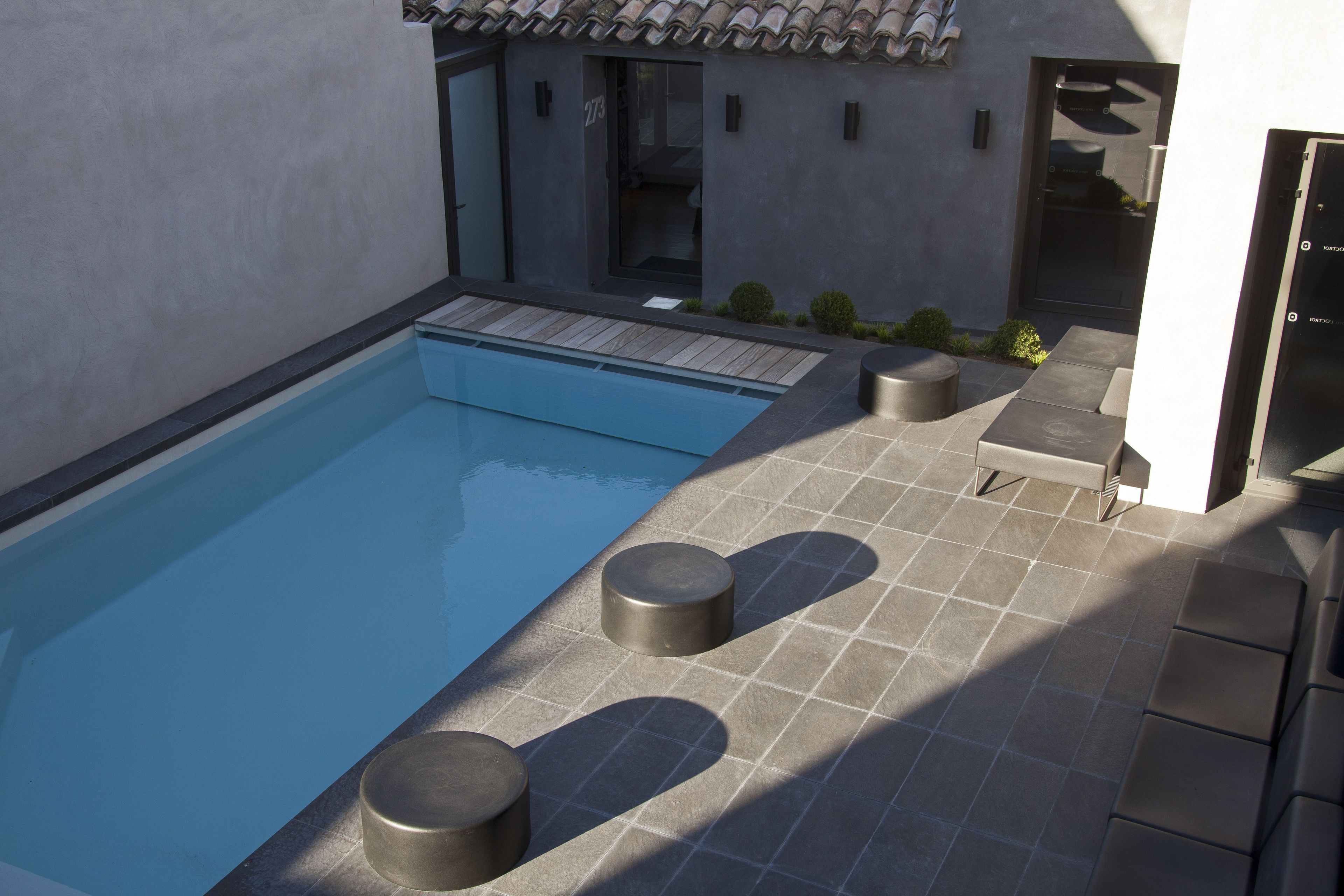 A modern outdoor pool area with a rectangular pool, stone tiles, and two cylindrical stools.