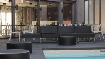 Un espace extérieur avec piscine, comprenant un coin salon avec un canapé et des fauteuils, ainsi qu’un bar visible à travers les portes vitrées.