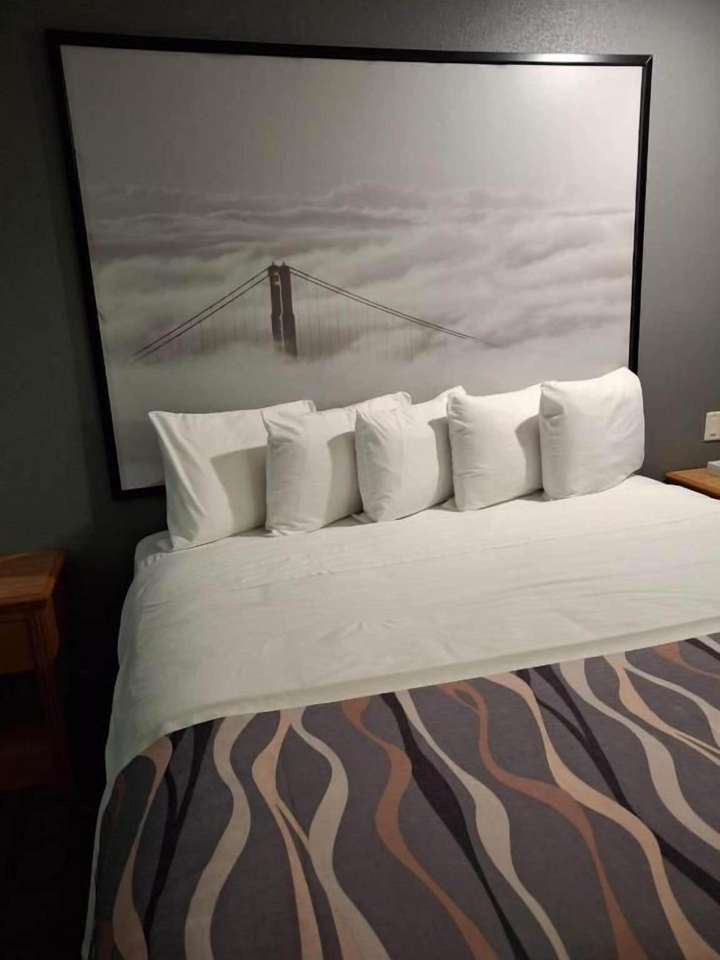 Un lit avec une literie blanche et une couverture à motifs, un grand tableau encadré représentant un pont accroché au mur, et une table de chevet en bois sur le côté gauche.