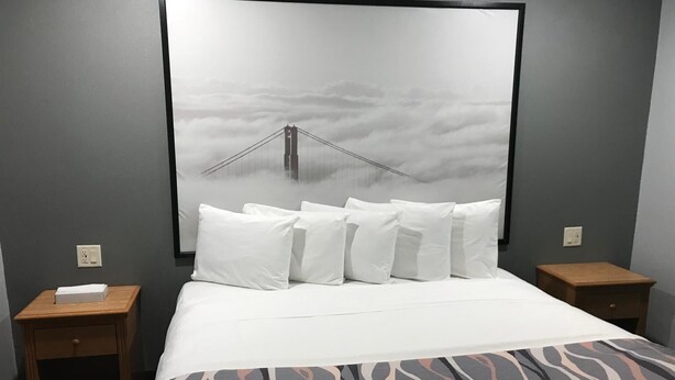 Une chambre d’hôtel avec un lit, des oreillers blancs, une couvre-lit à motifs, un grand tableau encadré représentant un pont, et deux tables de chevet.