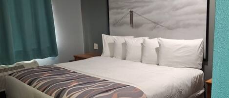 A hotel room with a large bed, a headboard with a framed picture, a window with curtains, and a wall-mounted air conditioner.