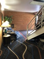 Un vestíbulo de hotel con una pared de ladrillo, un sofá con cojines, una lámpara, una alfombra con líneas onduladas y un letrero de salida.