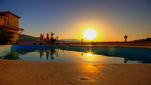 A swimming pool reflecting the sunset with a mountain backdrop and a building on the left.