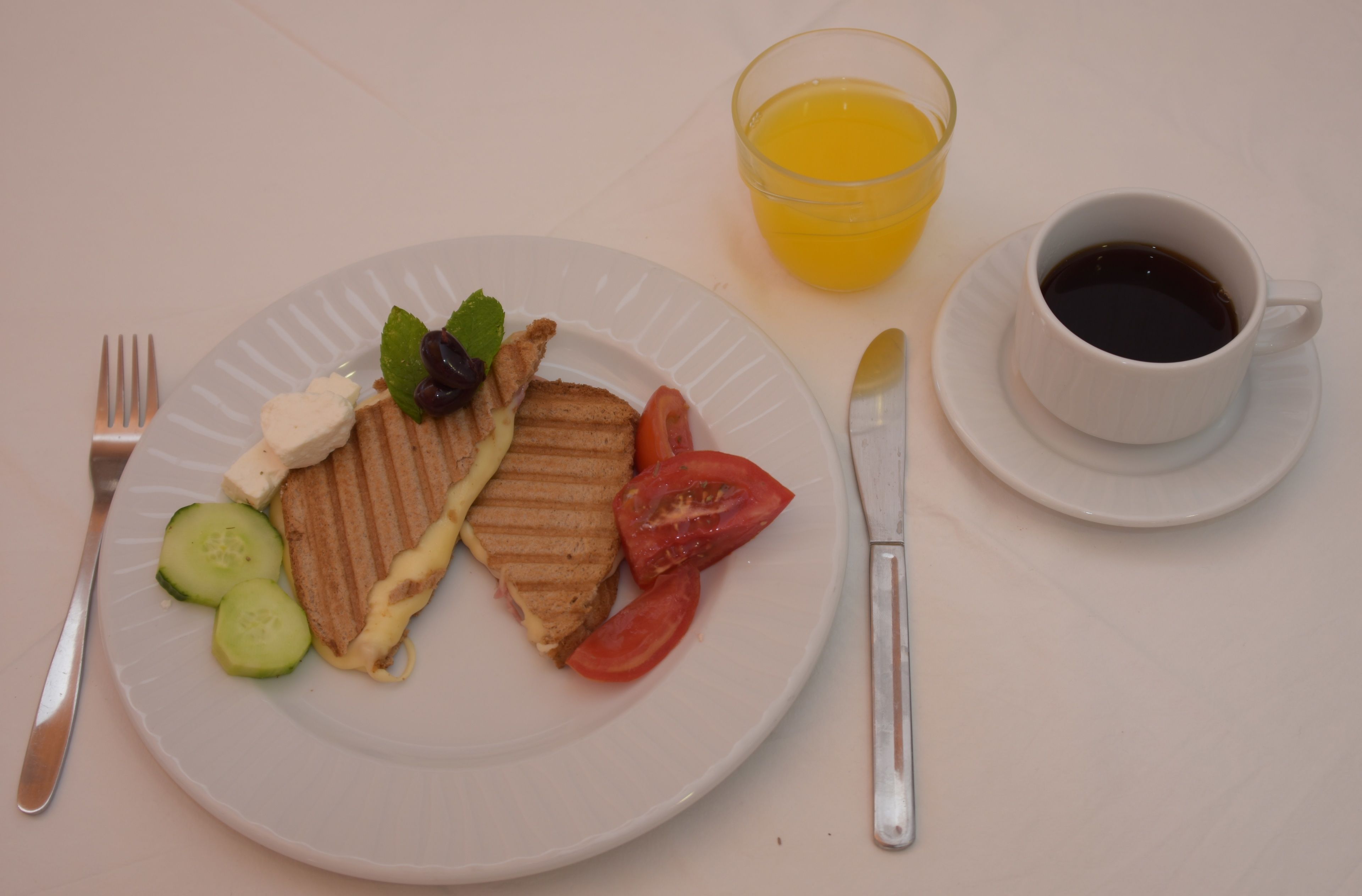 Een bord met een gegrilde kaas sandwich, tomaat, komkommer en een kleine salade. Op tafel staat een glas sinaasappelsap en een kop zwarte koffie.