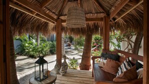 Une terrasse en bois surmontée d’un toit de chaume, un banc avec des coussins et une lanterne.