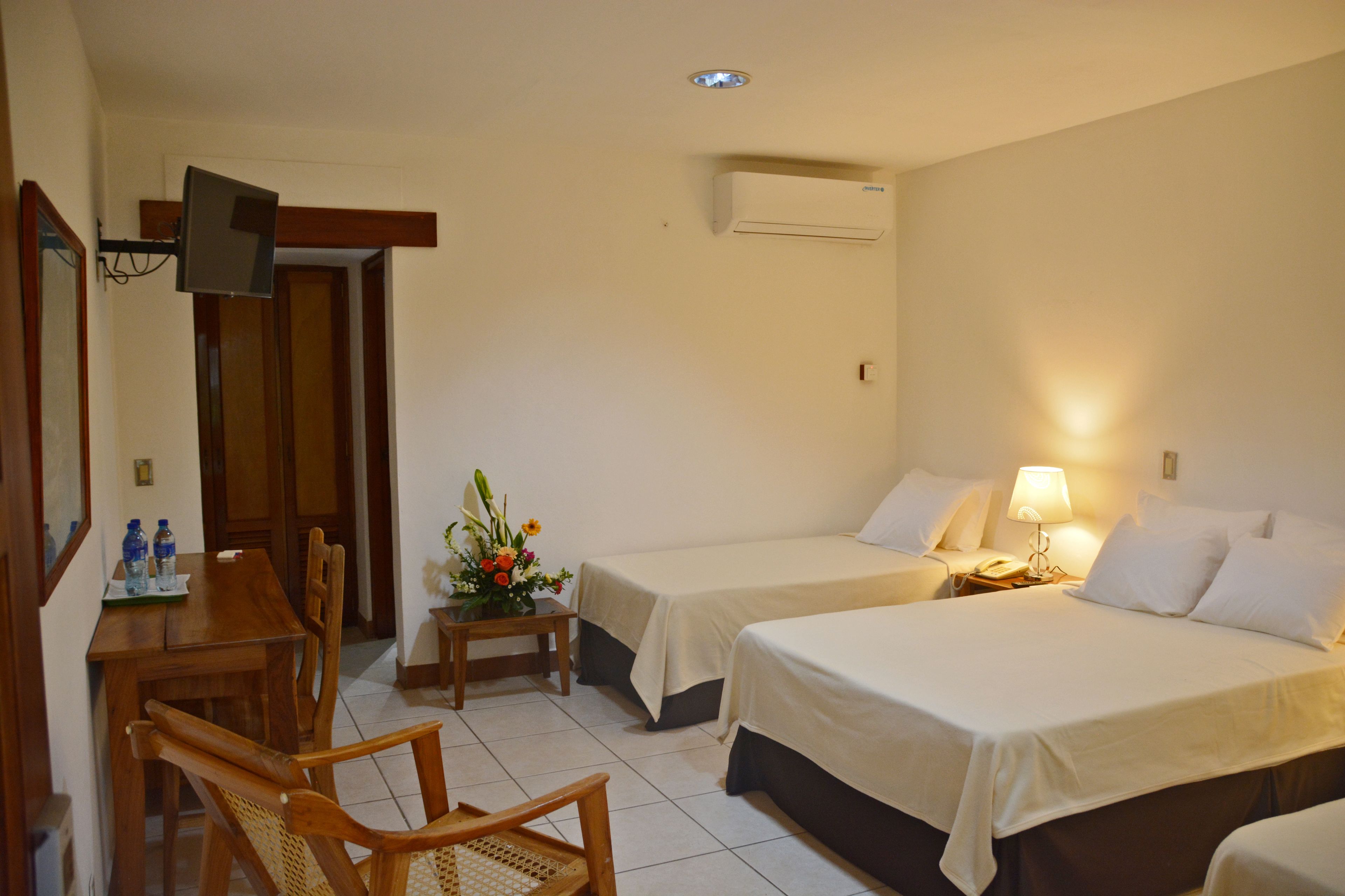 A hotel room with two beds, a desk with chairs, a TV, and a small table with a flower arrangement.