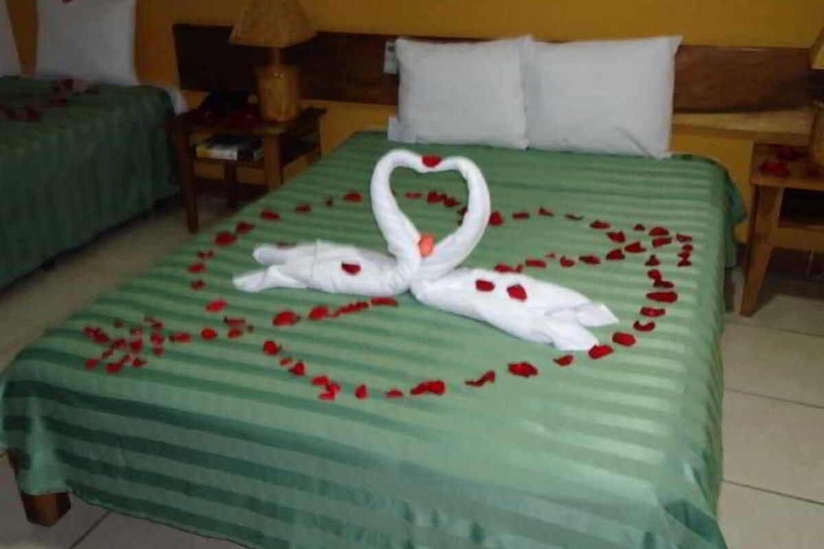 A hotel room with a bed covered in a green striped bedspread, a heart-shaped arrangement of rose petals, and two swan-shaped towels.