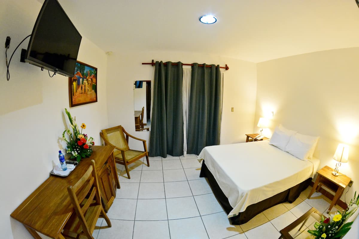 A hotel room with a bed, a desk with a chair, a TV mounted on the wall, and a painting on the wall.