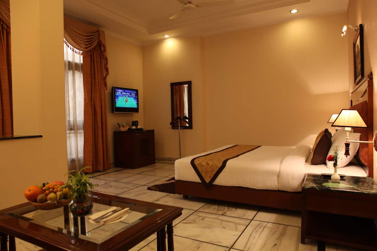 A hotel room with a bed, a TV, a desk with a lamp, a vase with flowers, and a fruit bowl.