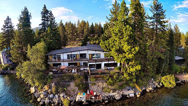 Un chalet à deux étages doté d’un balcon, entouré d’arbres et bordé par un lac.