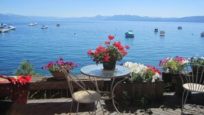 Une terrasse au bord du lac, avec une table ronde et des chaises, entourée de plantes en pot fleuries. On aperçoit des bateaux sur l’eau, au loin.