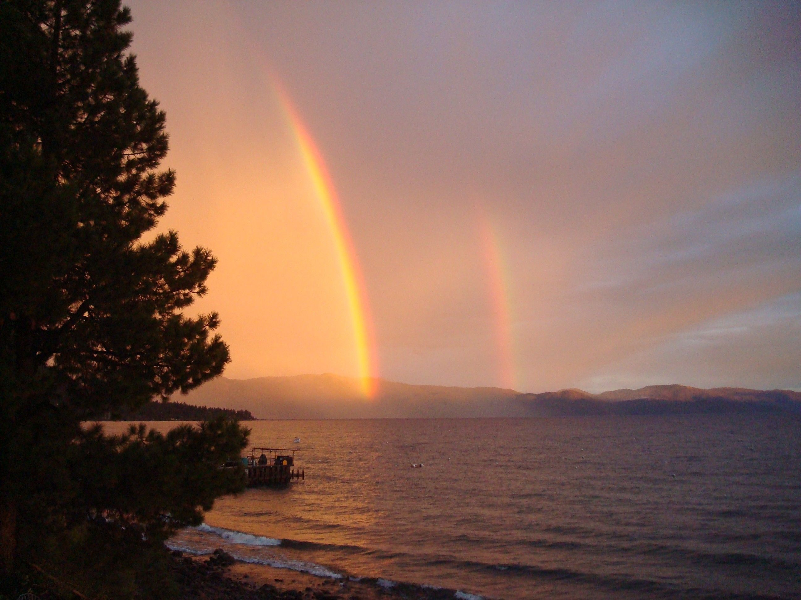 Eine Küstenlandschaft mit Regenbogen über dem Meer, einer Anlegestelle mit einem Boot und einem bewaldeten Gebiet.