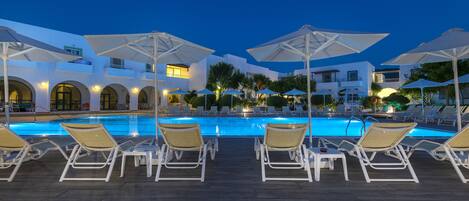 Zona junto a la piscina con sillones de descanso y sombrillas, piscina y un edificio al fondo con ventanas de arco y balcones.