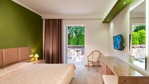 A hotel room with a bed, a desk, a chair, a TV, and a balcony with outdoor furniture. - Alexandra Beach Hotel (Kos)