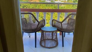 A balcony with wicker chairs and a small round table, overlooking a hillside with houses and greenery. - Hotel Alexandrina (Ribishi)