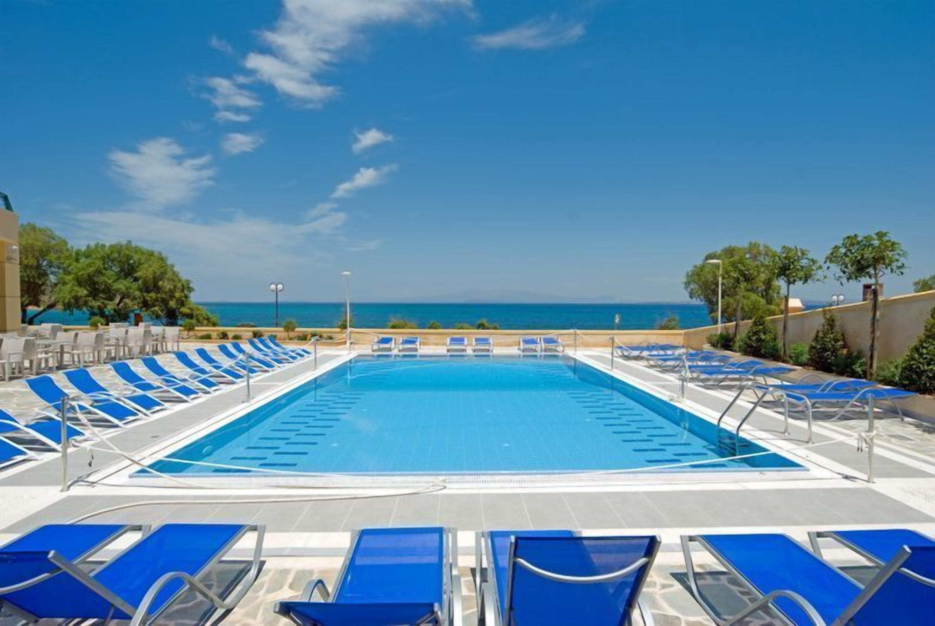 A rectangular swimming pool with blue water, surrounded by blue lounge chairs and a clear view of the sea.