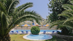 A pool area with lounge chairs and umbrellas, surrounded by palm trees and a stone wall. - Kyveli Apartments (Chios)