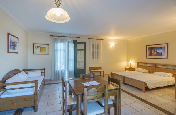 A room with two single beds, a wooden dining table, chairs, a bench, and a small table with a lamp. - Kyveli Apartments (Chios)