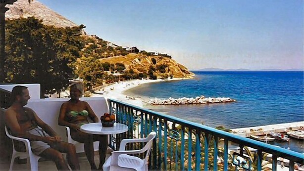 A seaside view with a rocky cliff, a beach, and a small pier with boats. - Kyveli Apartments (Chios)