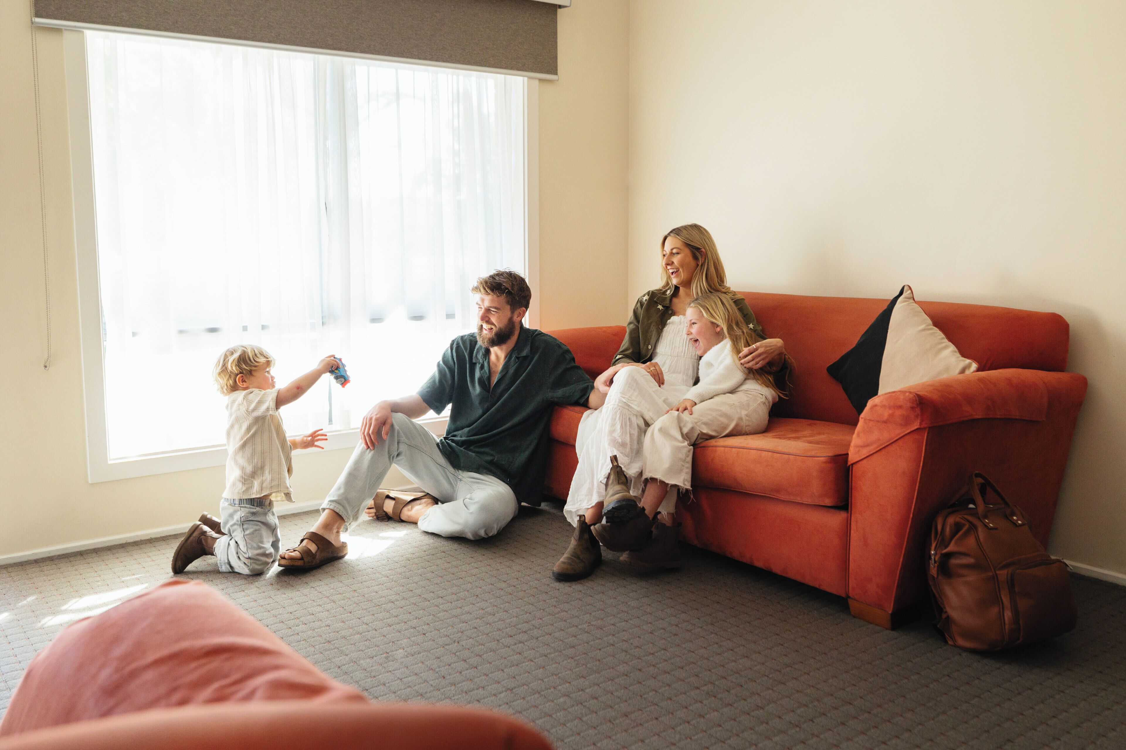 A family sitting on a couch in a room with a large window, carpeted floor, and a bag on the floor.