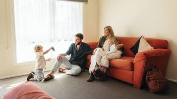 A family sitting on a couch in a room with a large window, carpeted floor, and a bag on the floor.