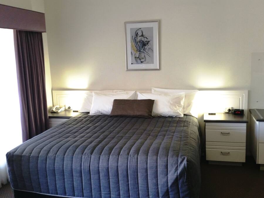 Ein Hotelzimmer mit einem großen Bett, zwei Nachttischen mit Lampen, einem gerahmten Kunstwerk an der Wand und einem Fenster mit Vorhängen.