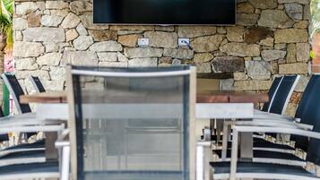 An outdoor patio area with stone walls, a flat-screen TV mounted on the wall, and multiple dining tables and chairs.