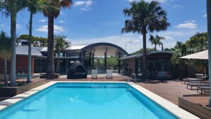 A swimming pool with lounge chairs and umbrellas, surrounded by palm trees and a covered seating area. - McLaren Vale Motel & Apartments (McLaren Vale)